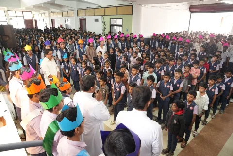 A large group of students in school uniform, many wearing coloured paper crowns, with a man in white in the foreground
